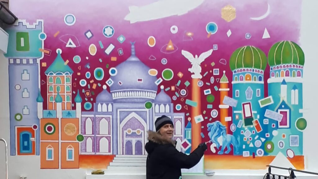 Stefan Eichler vor seiner Wandmalerei mit Kuppeln, Türmen, blauem Löwen und Zeppelin, gemalt für NewBoxes in Ravensburg