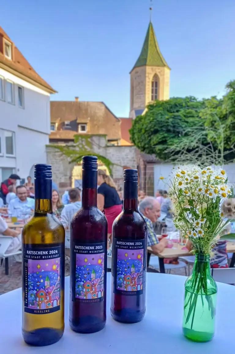 Drei Flaschen Ratsschenk der Stadt Weinstadt mit Etikett nach Stefan Eichlers Gemälde, im Hintergrund der Kirchturm aus dem Bild und Gäste beim Ratstrunk